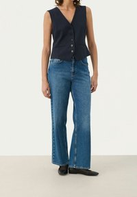 Marineblaue ärmellose Weste mit sechs Knöpfen und zwei Taschen, kombiniert mit hochgeschnittenen, weiten blauen Jeans und schwarzen flachen Schuhen.