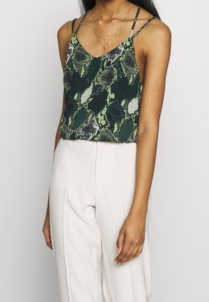 Woman wearing sleeveless green and black snake print top with thin straps and high-waisted white trousers, visible from shoulders to hips.
