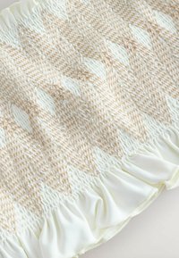 Krämfärgad topp med smockad yta, med ett zigzag-mönster i ljusbeige. Designen har en volangkant för extra detalj.