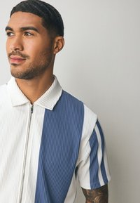 Kurzärmeliges Hemd mit Kragen, das blaue und weiße vertikale Streifen, eine gerippte Struktur und einen Reißverschluss vorne aufweist.