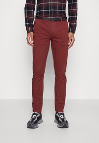 En person står klädd i en svart och röd rutigt skjorta, med åtsittande röda byxor och svarta sneakers, framför en enkel vit bakgrund.