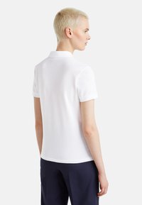 Weißes Polohemd mit Kragen und kurzen Ärmeln, glatte Textur, getragen mit einer Marineblauen Hose. Von hinten betrachtet, zeigt es minimale Designdetails.