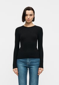 Γυναίκα με κοντά σκούρα μαλλιά, που φοράει εφαρμοστό μακρυμάνικο μαύρο τοπ και μπλε τζιν, στέκεται μπροστά σε απλό λευκό φόντο.