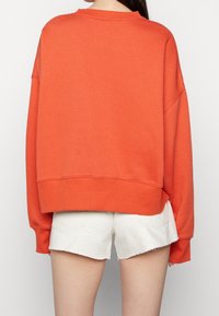 Pomarańczowa, oversize'owa bluza z szerokimi mankietami i okrągłym dekoltem, zestawiona z białymi, przetartymi szortami z postrzępionymi brzegami.