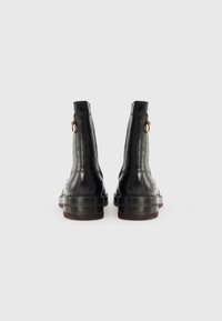 Botas de cano curto em couro preto com uma textura lisa, apresentando um detalhe em zíper dourado na parte de trás e uma sola grossa e transparente.