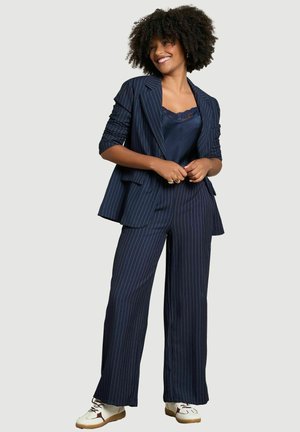 Femme aux cheveux bouclés portant un costume à fines rayures bleu marine, un top en dentelle bleu foncé et des baskets blanches, souriante, les mains jointes devant elle.