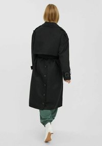 Czarny trench z opadającymi ramionami, dużym zakładanym tyłem i zapięciem na guziki. Noszony na zielonych dresach i beżowych botkach na obcasie.