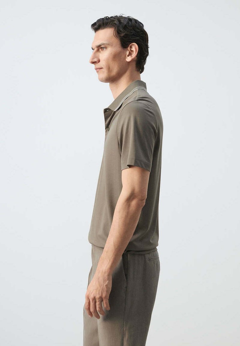 Man standing in profile wearing a taupe short-sleeve polo shirt and matching trousers against a plain light background.