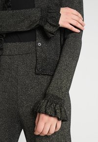 Zwart en goud gebreide top met gerimpelde manchetten, voorzien van een knoopsluiting en gestructureerde stof. De outfit omvat nauwsluitende broek met een gladde afwerking.