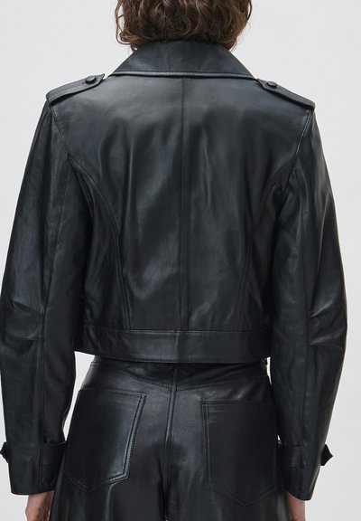 Άτομο που φοράει μαύρο δερμάτινο κοντό μπουφάν και ταιριαστό δερμάτινο παντελόνι, εμφανιζόμενο από πίσω με φόντο ένα απλό ανοιχτό φόντο.