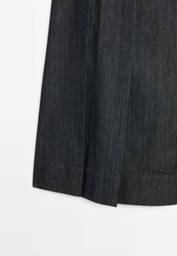 Détail d'un tissu en denim gris foncé montrant l'ourlet et les coutures verticales d'une jupe ou d’un pantalon sur fond blanc.