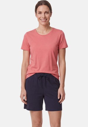 Vrouw staand, glimlachend, draagt een koraalroze T-shirt met korte mouwen en marineblauwe shorts met trekkoord tegen een effen lichte achtergrond.