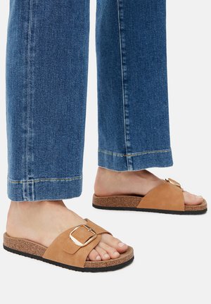 Zamszowe klapki w kolorze jasnobrązowym z szerokim paskiem i złotym klamrą, uzupełnione korkową podeszwą i czarnym gumowym spodem.