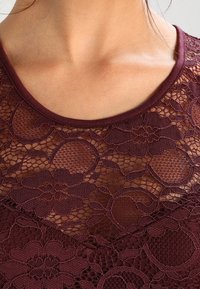 Kobieta w burgundowej koronkowej bluzce w kwiaty, z przejrzystymi i nieprzezroczystymi sekcjami materiału oraz okrągłym dekoltem.
