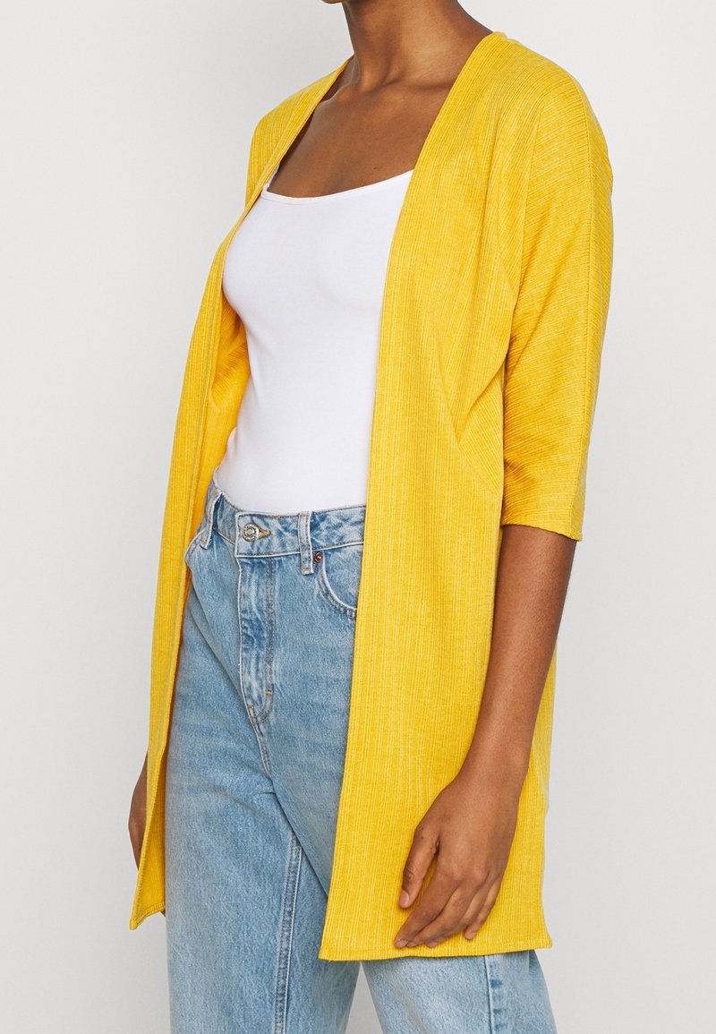 Gul stickad kofta med öppen front, mittlång ärm och avslappnad passform, kombinerad med en vit linne och ljusblå jeans.
