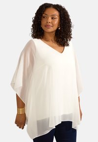 Vit blus med v-ringning gjord av lätt, semi-transparent tyg. Har flödande ärmar och en rundad nederkant, i kombination med mörka byxor.