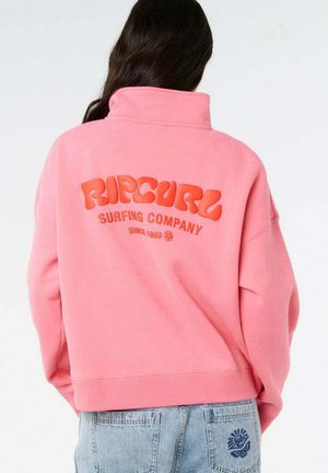 Persona con capelli lunghi e scuri che indossa una felpa rosa Rip Curl Surfing Company e jeans azzurri con ricamo blu sulla tasca posteriore.