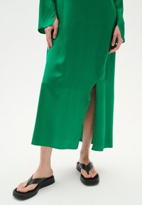 Zöld selyem maxi ruha oldalsó hasítékkal, fekete flip-flop szandálokkal párosítva, vastag talppal és minimalista kialakítással.