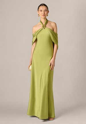 Femme portant une longue robe col halter vert olive avec manches tombantes, boucles d'oreilles dorées, tenant une petite pochette dorée.