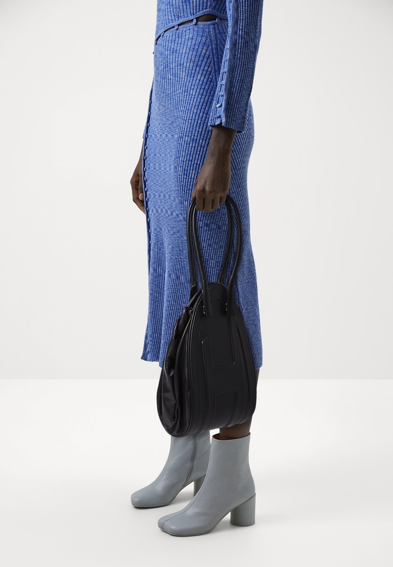 Femme portant une robe côtelée bleue et des bottines gris clair, tenant un sac à main en cuir noir par ses anses à son côté.