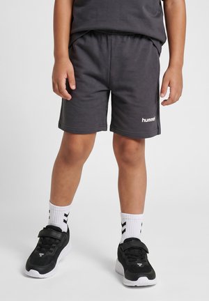Kind draagt zwarte sportshorts, zwarte sneakers met witte zolen en witte sokken met zwarte chevronpatronen, staand op een grijze vloer.