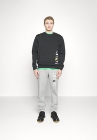 Felpa nera con diversi logo Nike, pantaloni soft grey con logo e scarpe nere. Capelli corti e ricci, in piedi davanti a uno sfondo neutro.