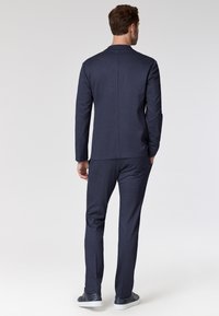 Completo due pezzi blu navy in un tessuto testurizzato, con giacca a maniche lunghe dal taglio dritto e pantaloni su misura, abbinato a sneaker.