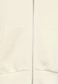 Sudadera crema con cremallera central, dobladillo acanalado y gran bolsillo frontal. Textura suave con un diseño sin costuras y sin patrones visibles.