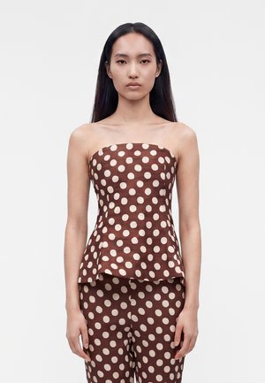 Femme aux longs cheveux noirs portant un haut marron sans bretelles et un pantalon à gros pois blancs, debout devant un fond blanc uni.