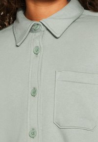 Camicia verde chiaro con bottoni, caratterizzata da un colletto classico, tessuto testurizzato, quattro bottoni e una tasca sul petto a sinistra.
