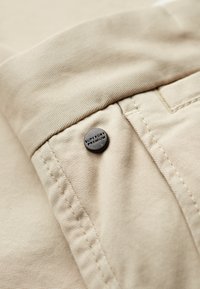 Beżowa tkanina bawełniana z widocznym szwem i czarnym guzikiem z napisem "SUPERDRY PREMIUM" przymocowanym do pasa.