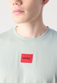 Svetlozelené bavlnené tričko s okrúhlym výstrihom a červeným obdĺžnikovým nášivkom s nápisom "HUGO" v čiernej farbe. Hladká textúra, bez vzorov.