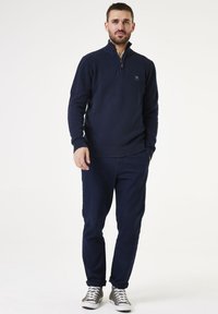 Marineblauer Strick-Half-Zip-Pullover mit strukturiertem Muster, gerippten Bündchen und Saum, kombiniert mit marineblauen Chinos und grauen Sneakern, vor einem weißen Hintergrund stehend.