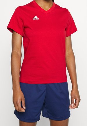 Person iført en rød Adidas T-shirt med korte ærmer og V-hals samt mørkeblå sportsshorts, stående mod en ensfarvet baggrund.