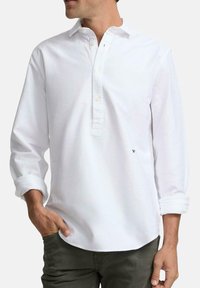 Camisa blanca de manga larga con tapeta y detalle de botones. Presenta un corte holgado y puños enrollados. Destaca un pequeño logo cerca del dobladillo.