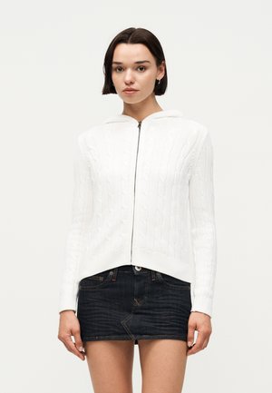 Γυναίκα με κοντά σκούρα μαλλιά που φοράει λευκή ζακέτα με φερμουάρ και πλεκτό σχέδιο καλωδίου και μαύρη μίνι φούστα από τζιν, στέκεται μπροστά σε ένα απλό φόντο.