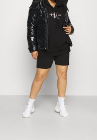 Giacca a vento nera lucida, felpa con cappuccio nera con logo, pantaloni corti neri, sneakers bianche con dettagli grigi, calzini bianchi alla caviglia.