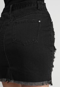 Černá džínsová sukně s roztrhaným lemem, vybavená dvěma zadními kapsami a jemným opotřebením. Materiál je strukturovaný s klasickým střihem.