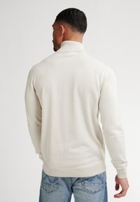 Suéter de cuello alto en crema claro, hecho de una tela suave, con un diseño ajustado y puños acanalados. Lleva puesto unos jeans azules.