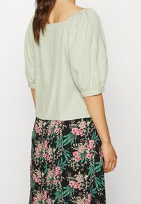 Femme portant un chemisier à manches bouffantes vert clair et une jupe noire avec des motifs floraux roses et verts, vue de dos.