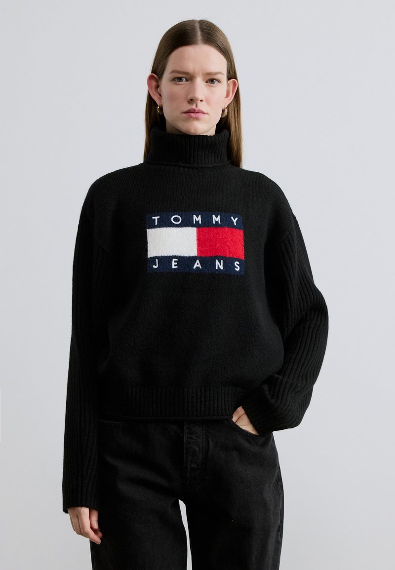 Pull à col roulé en maille noire avec manches et ourlet côtelés, arborant un patch central avec un logo en blocs bleu, blanc et rouge.