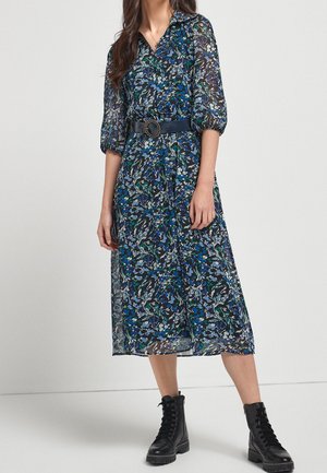 Robe midi florale aux tons bleus et verts, dotée d'un col à revers, de manches bouffantes et d'une ceinture en tissu avec une boucle circulaire. Portée avec des bottines noires à lacets.