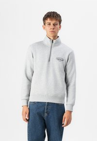Grå pullover med høj krave og halv lynlås, der har et logo på brystet. Fremstillet af blødt materiale, kombineret med blå jeans.