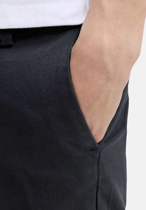 Main partiellement dans la poche avant d’un pantalon noir, montrant la texture du tissu et les détails des coutures.