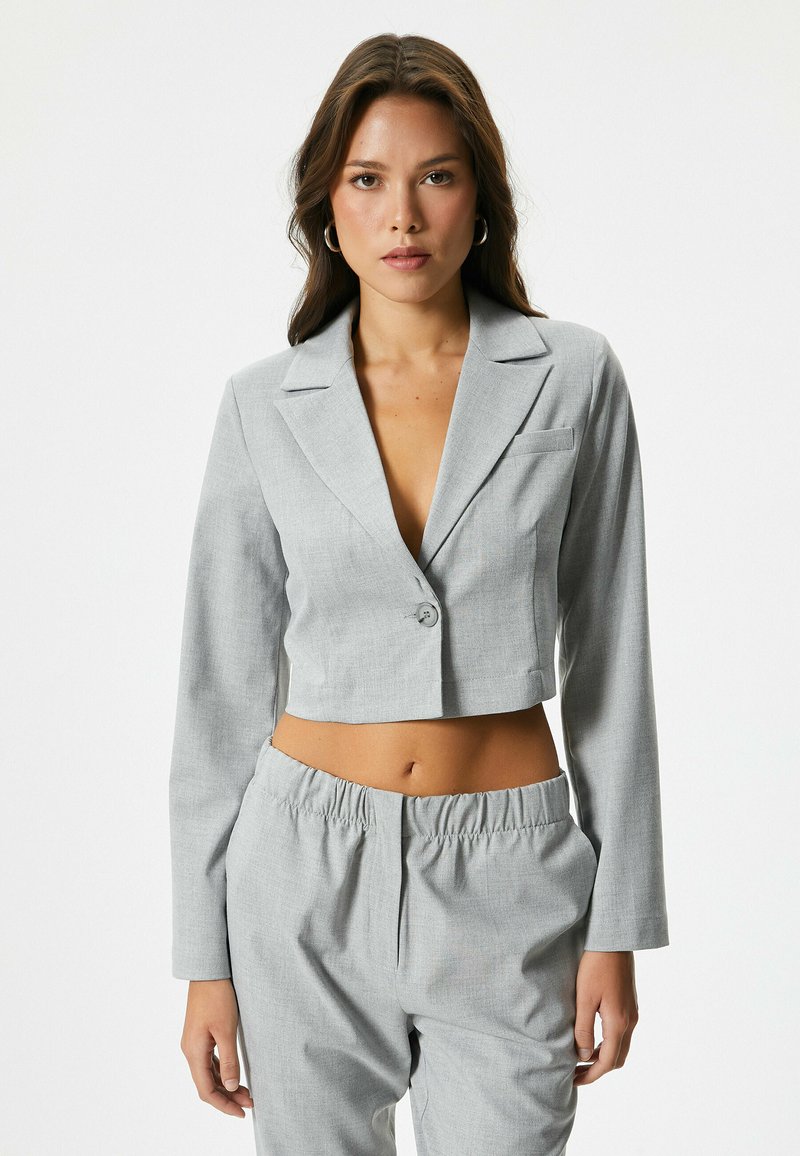 Blazer gris corto con mangas largas, solapas de pico, cierre de un solo botón y un pequeño bolsillo frontal. Combinado con pantalones ajustados a juego.