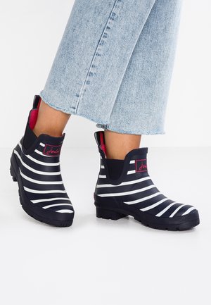 Nilkkaan ylettyvät mustavalkoraidalliset kumisaappaat, joissa on vaaleanpunainen vuori, yhdistettynä lyhytlahkeisiin vaaleansinisiin farkkuihin.