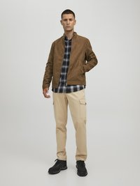 Giacca scamosciata marrone con colletto alto, camicia a quadri bianca e nera sotto, pantaloni cargo beige e scarpe nere.