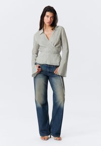 Blouse enveloppante gris clair en tissu texturé avec des manches larges, associée à un jean droit bleu délavé et des sandales ouvertes beige.