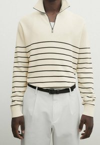 Pull crème à rayures horizontales noires, col zippé, coupe ajustée, assorti à un pantalon gris clair et une ceinture noire.