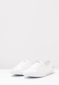 Baskets en toile blanches avec un design basse, bout rond, fermeture à lacets et semelle en caoutchouc. Marquage minimal sur la languette et le côté.